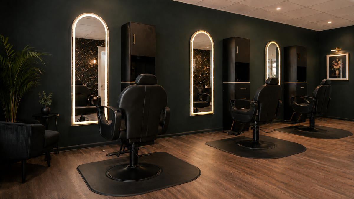 A modern barbershop or salon interior with four black barber chairs arranged in front of tall arched mirrors with warm white lighting, set in a room with dark green walls and wood flooring.